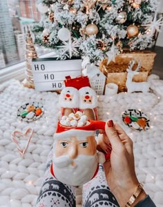 someone holding up a santa clause mug in front of a christmas tree with other decorations