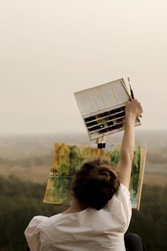 a woman is holding up an open book with paintbrushes in her left hand