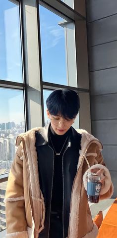 a young man standing in front of a window holding a cup and looking at his cell phone