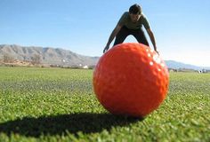 a man bending over to pick up an orange ball on the grass with mountains in the background