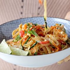 a person is pouring sauce on a bowl of noodles with vegetables and lime wedges