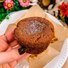 a hand holding a chocolate muffin on top of a white plate with flowers in the background