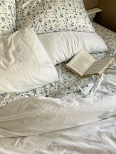 an open book sitting on top of a bed with white sheets and pillows in front of it