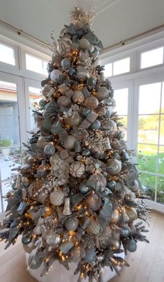 a decorated christmas tree with silver ornaments and lights in a room next to large windows