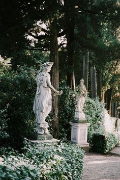 two statues in the middle of a garden surrounded by bushes and trees, one holding a woman's hand