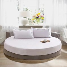 a white round bed with pillows on it in front of a window and vase filled with flowers