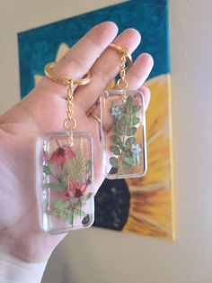 two clear keychains with flowers and leaves on them in front of a painting