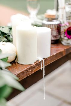 two white candles sitting on top of a wooden table next to flowers and greenery