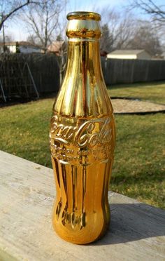 a gold coca - cola bottle sitting on top of a wooden table in front of a yard