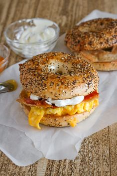 two bagels with eggs, bacon and cheese on them sitting on wax paper next to a bowl of yogurt