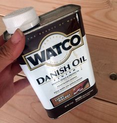 a hand holding onto a bottle of watch danish oil on a wooden table with wood grains in the background