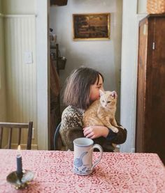 a woman sitting at a table with a cat on her lap and a coffee mug in front of her