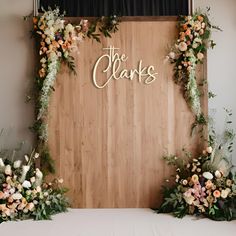 a wooden wall with flowers and greenery on it that says the clarks in gold lettering