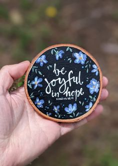 a hand holding a small black and blue flowered coaster that says be joyful in hope