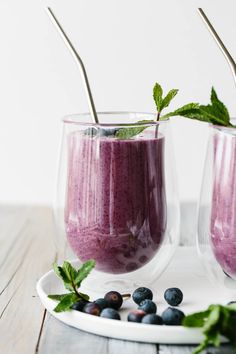 two glasses filled with blueberry smoothie on top of a white plate next to berries