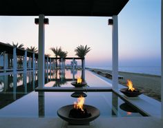 an outdoor fire pit on the side of a swimming pool at dusk with palm trees in the background