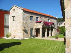 an old stone house with red flowers on the front and side windows, along with green grass