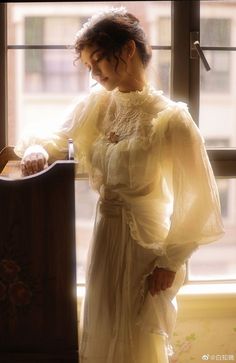 a woman standing in front of a window wearing a white dress and holding onto a wooden chair