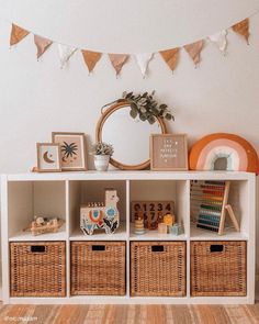 a white shelf with baskets and pictures on it next to a mirror, potted plant and other items