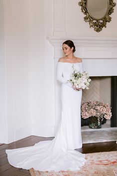 a woman standing in front of a fire place holding a bouquet of flowers and wearing a white dress