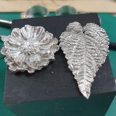 two silver leaf shaped brooches sitting on top of a black piece of paper