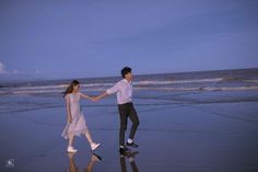 a man and woman holding hands while walking on the beach at dusk with waves crashing in