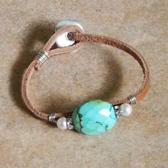 a leather bracelet with a turquoise bead and white pearls on it, sitting on a brown surface