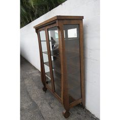 a wooden display case with glass doors on the side of a white wall next to a tree