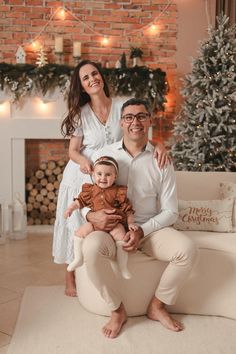 a man and woman holding a baby in front of a fireplace with christmas lights on it