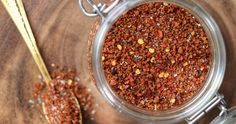 a glass jar filled with red chili seasoning next to two spoons on a wooden surface