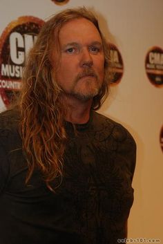 a man with long hair standing in front of a red carpeted wall and wearing a black shirt