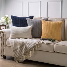 a couch with four different colored pillows on it and a vase full of flowers in the corner