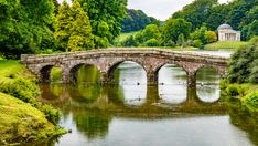 a stone bridge over a body of water surrounded by lush green trees and greenery