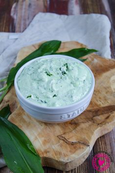 a white bowl filled with green cream sitting on top of a wooden cutting board next to leaves