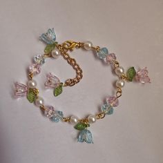 This bracelet is 7.5 inches long with a 2-inch extender. It has pink and blue flower charms with green leaf accents, clear beads, and faux pearls, all connected by a gold chain. It fastens with a lobster clasp. Floral Beaded Bracelet, Fairycore Bracelet, Fairy Bracelet, Fairy Bracelets, Clear Bracelet, Green Beaded Bracelets, Bracelet Sets, Clear Beads, Pink And Blue Flowers