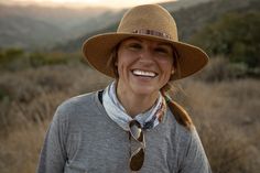 a woman wearing a hat and scarf standing in front of a field with mountains behind her