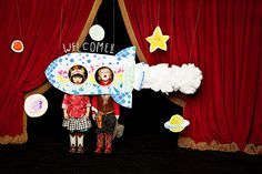 two children standing in front of a red curtain with a rocket ship cutout on it