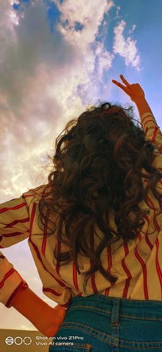 a woman with long hair standing in front of a blue sky holding her hands up