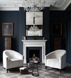 two white chairs sitting in front of a fireplace with a chandelier hanging from the ceiling