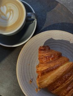 a croissant on a plate next to a cup of cappuccino