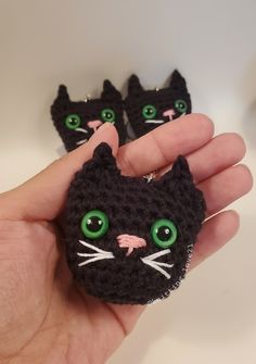 a hand holding two small black crocheted cat brooches with green eyes