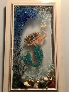 a glass frame with a mermaid and sea shells on the bottom is hanging on a wall