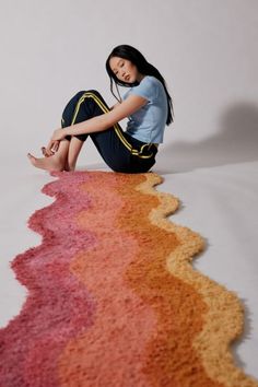 a woman sitting on the ground with her legs crossed and looking at the camera, in front of a multicolored rug