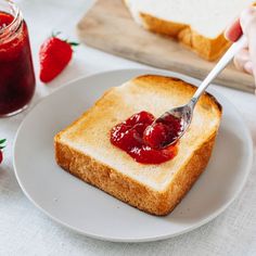 someone spooning jelly onto a piece of bread on a white plate with strawberries