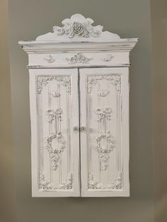 a white cabinet with ornate carvings on the doors and drawers is against a gray wall