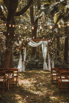 A beautiful garden wedding setup with a wooden arch draped in white fabric and adorned with floral arrangements, surrounded by string lights and wooden chairs under tall trees. Wedding Garden Ideas Outdoor Events, Forest Wedding Dinner, Magical Backyard Wedding, Cottagecore Backyard Wedding, Summer Cottage Wedding, Summer Outdoor Wedding Ideas On A Budget, Hobbit Wedding Aesthetic, Wedding Inspo Outdoor, Things To Have At Your Wedding