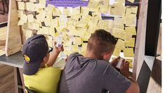 two boys sitting at a table with post it notes on the wall