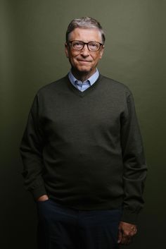 an older man wearing glasses standing in front of a dark green wall with his hands in his pockets