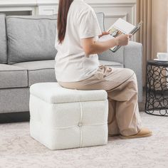 a woman sitting on top of a white ottoman next to a gray couch holding a book