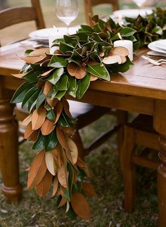 a wooden table topped with lots of greenery next to white plates and silverware